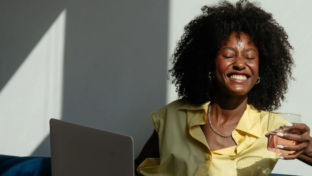 a woman in a yellow top smiling as she drinks pink coloured water from a glass with the sun shining in through the window.